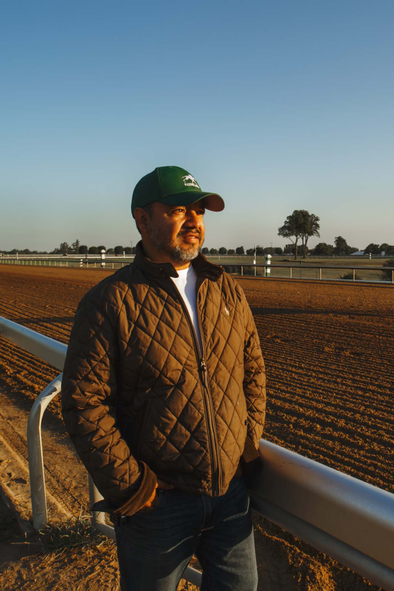 A photo of Alfredo Laureano looking off into the distance while standing by the dirt track at sunset. He is an older Hispanic man with graying chin stubble. He is wearing a green Keenland-brand hat and a brown Keenland-brand jacket overtop a white shirt.