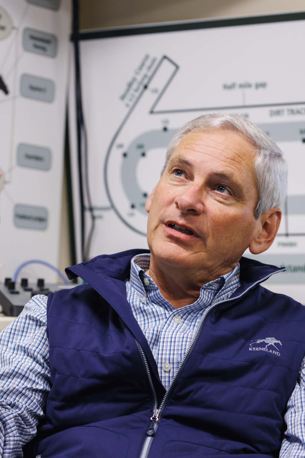 An up-close photo of Dr. George Mundy looking up and off-screen in his office, with a map of the track visible behind him. He is an older White man with short silver hair. He is wearing a navy Keenland-brand vest over a white and blue checkered shirt.